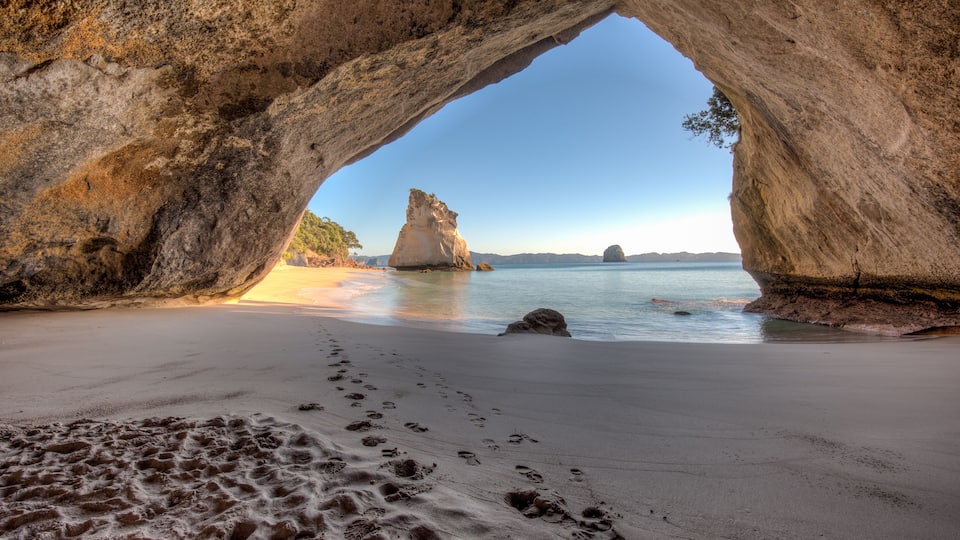 A natural rock arch formation over a beach with footprints leading to the water.