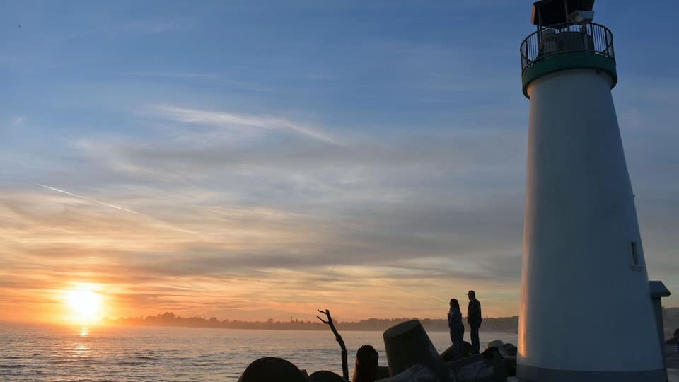 A lighthouse at sunset with silhouettes of people and a tree in the foreground.