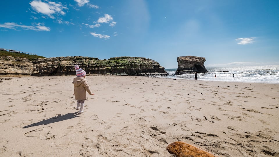A sandy beach with footprints, a person in a pink hat, and a large rock formation in the distance.