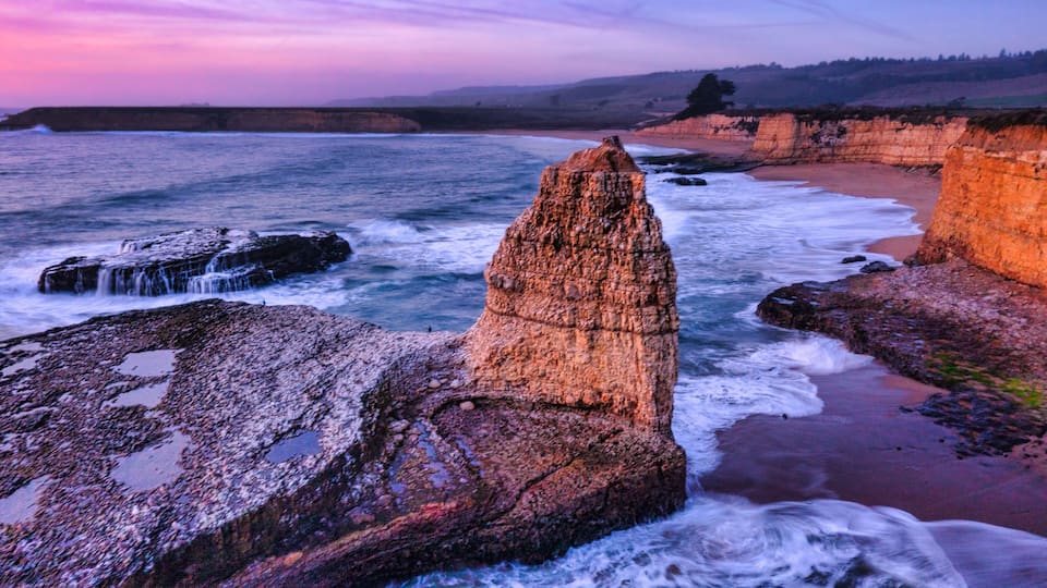 A coastal landscape with a prominent rock formation, waves crashing against the shore, and a sunset sky.