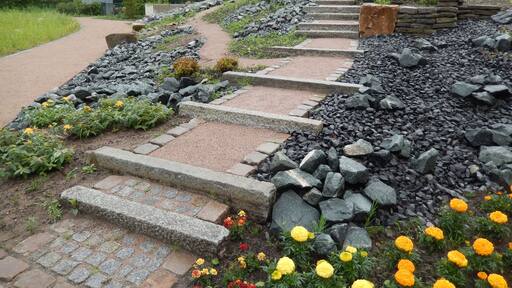 Un camino de piedra con escalones que ascienden por una pendiente, rodeado de rocas y plantas con flores.