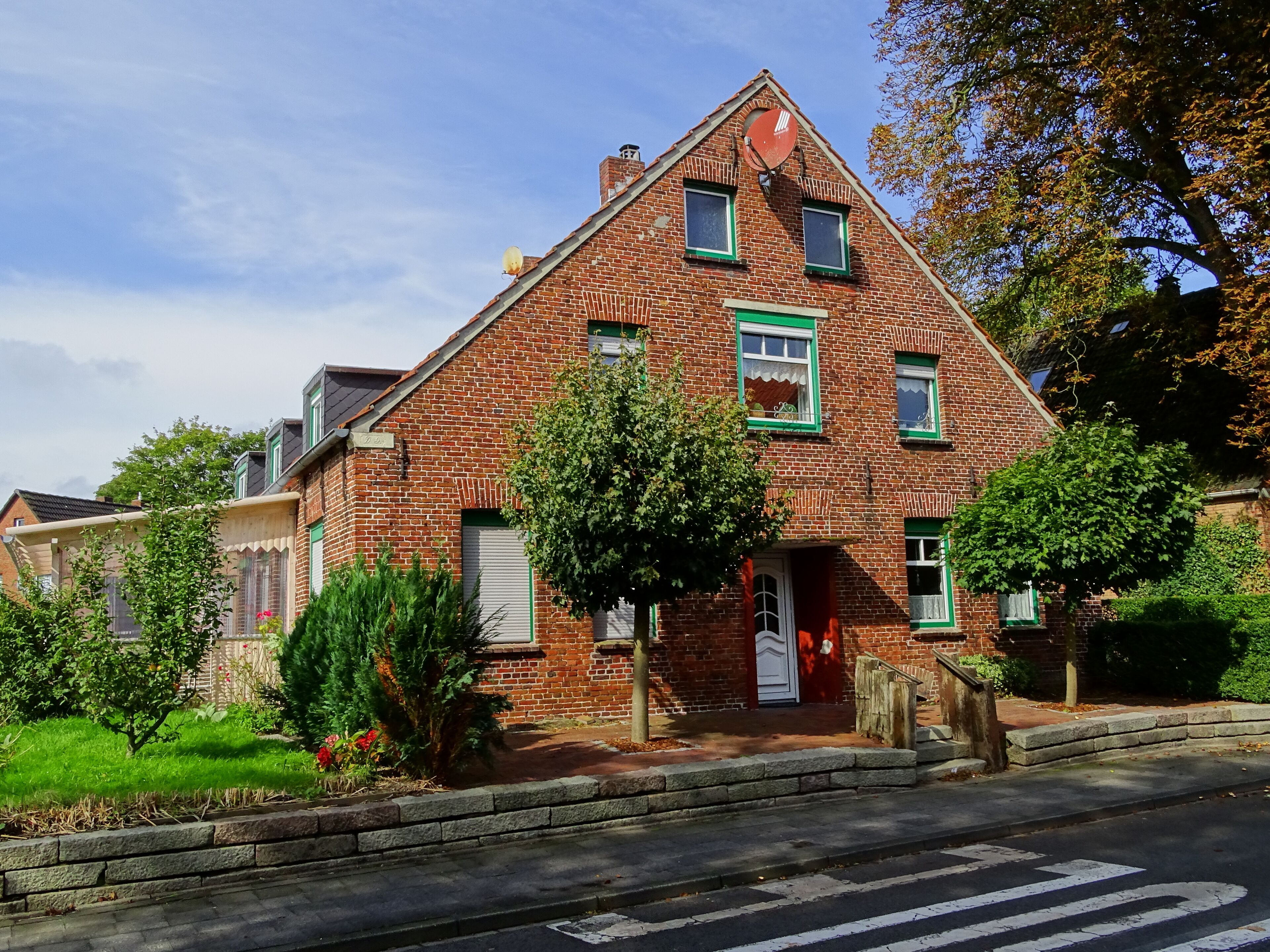 A red brick house with green trim, a white door, and a small garden with a tree and flowers.