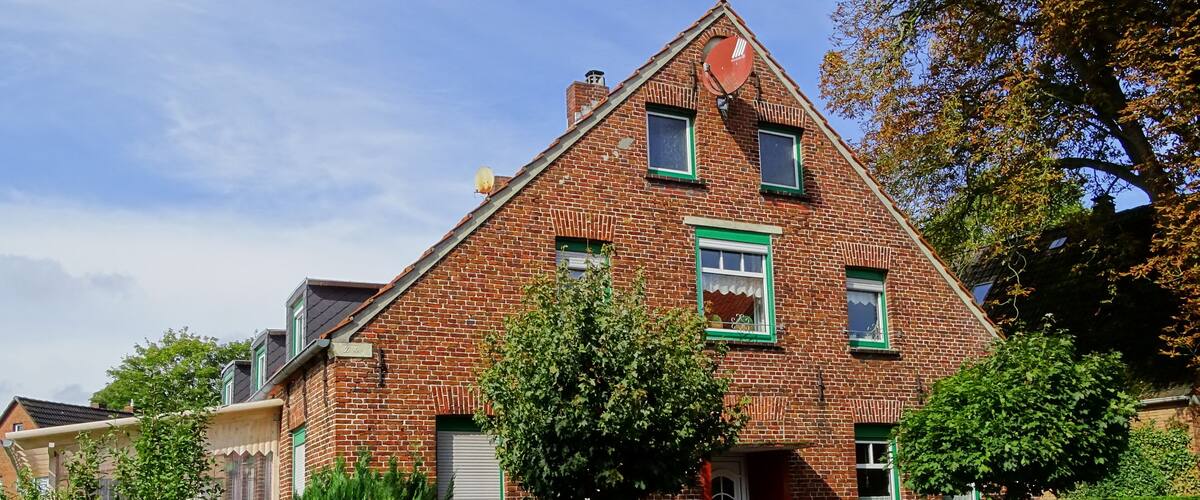 A red brick house with green trim, a white door, and a small garden with a tree and flowers.