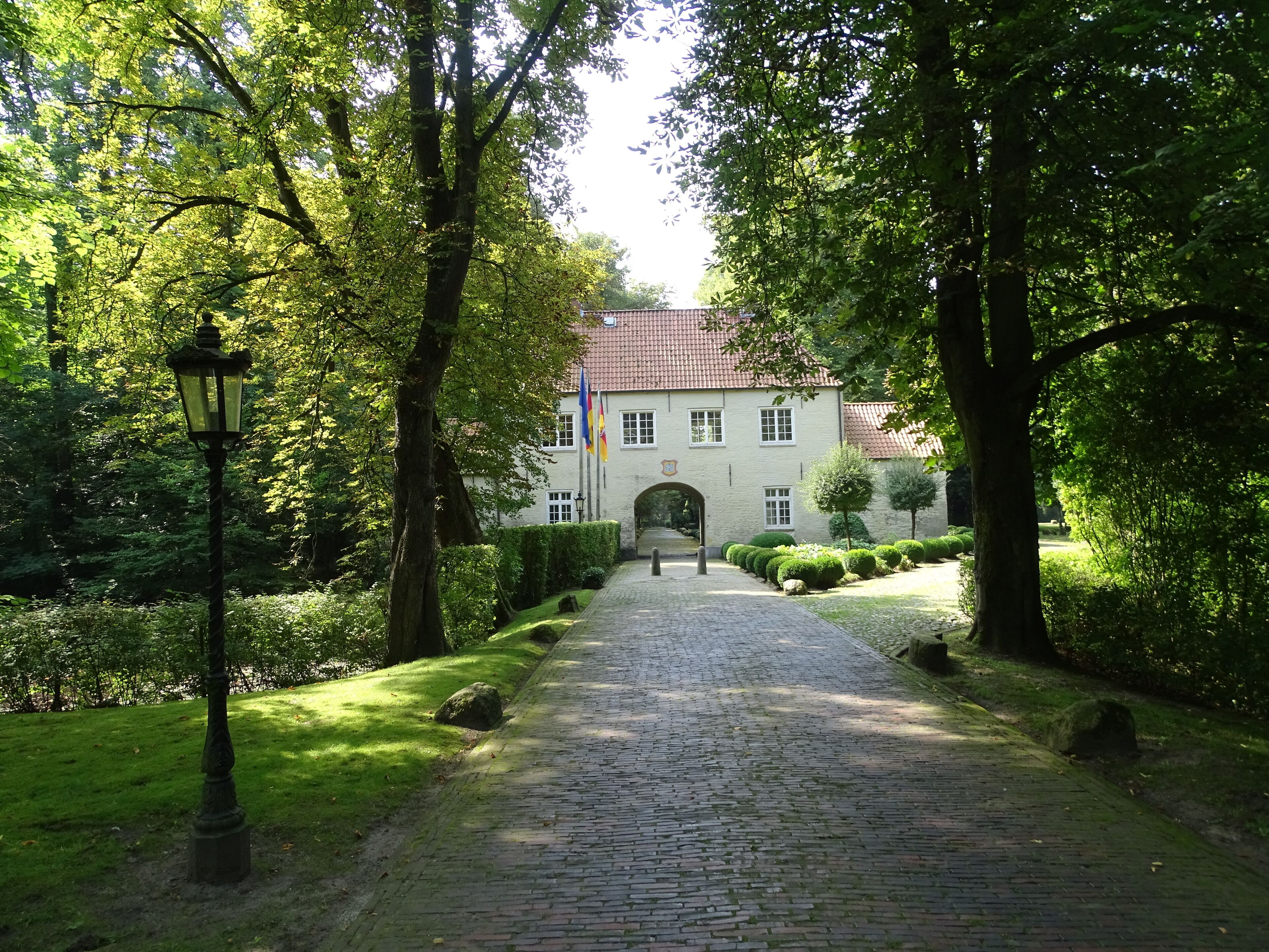 A cobblestone path leading to a building with a red-tiled roof, surrounded by lush greenery and a lamppost.