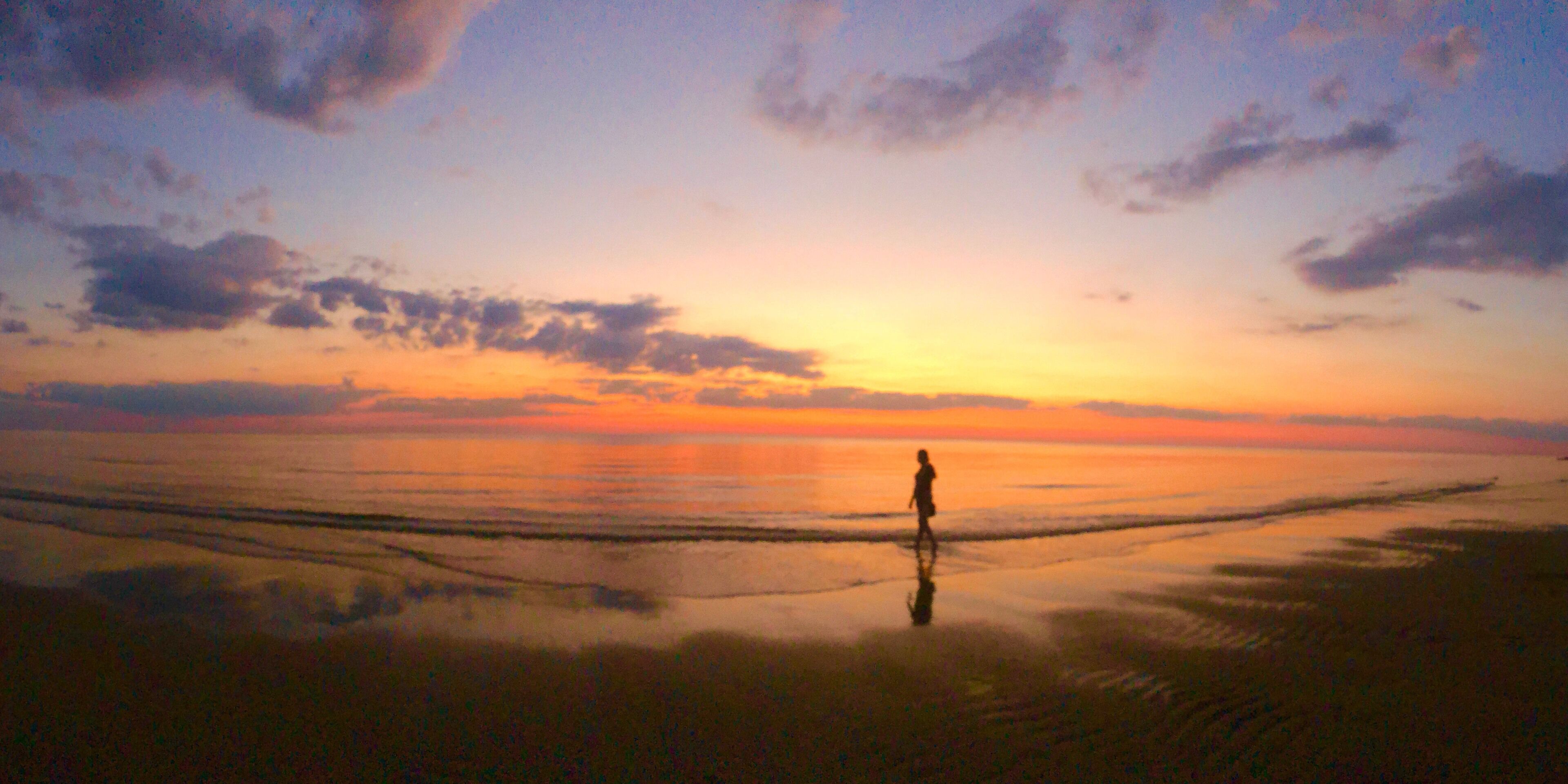 A person standing on a beach at sunset with a calm sea and a clear sky.