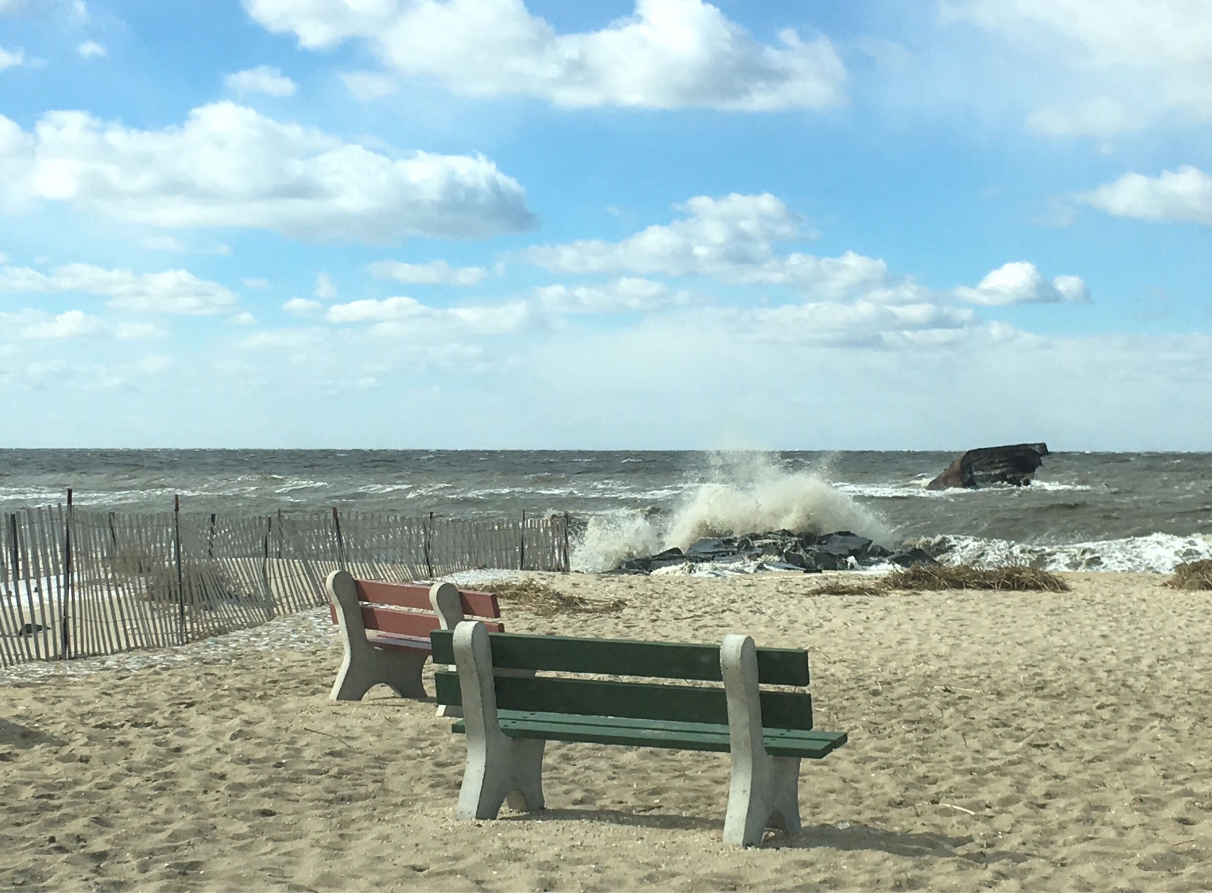 A beach with a wooden fence, a bench, and waves crashing against rocks.
