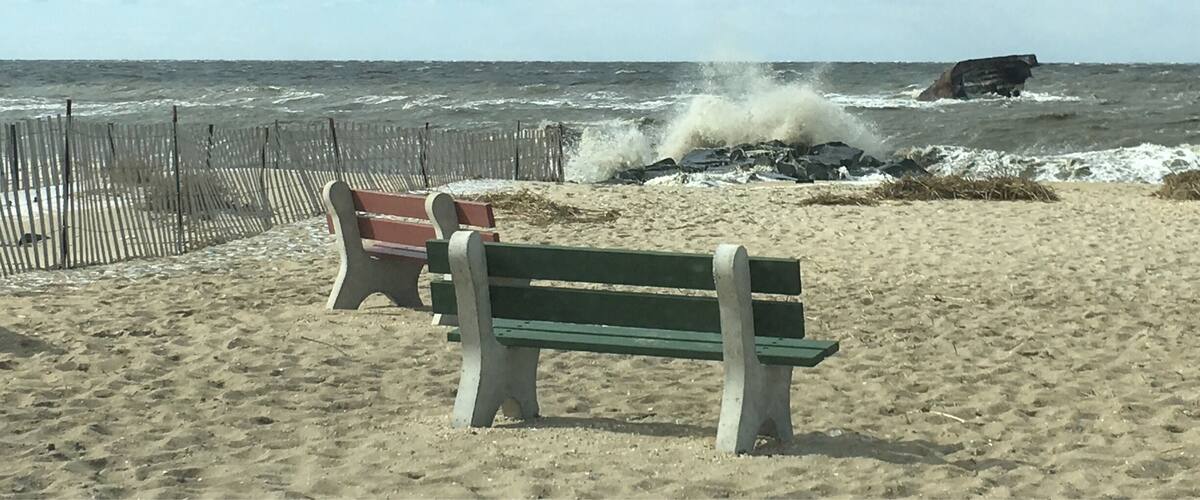 A beach with a wooden fence, a bench, and waves crashing against rocks.