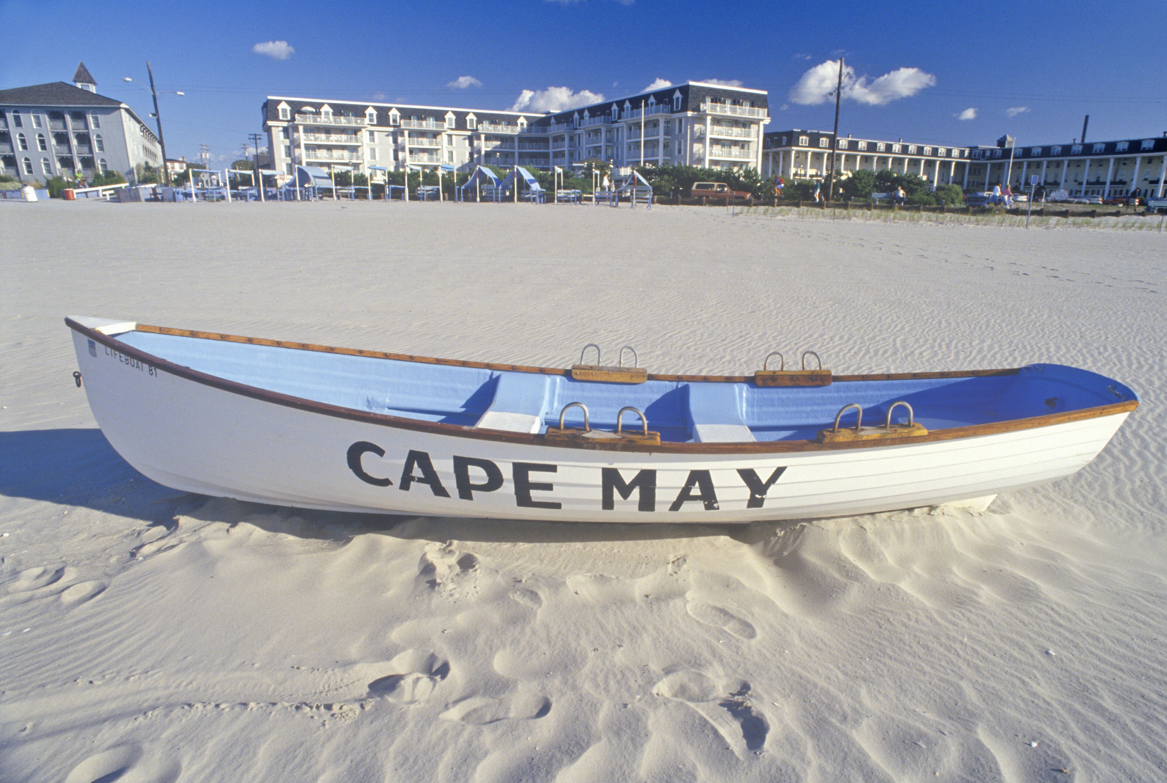 A white boat with 'CAPE MAY' written on it, parked on a sandy beach with buildings in the background.