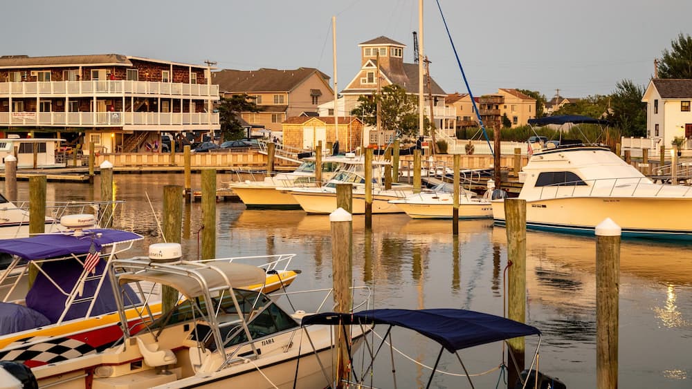 A marina with several boats docked, a wooden pier, and residential buildings in the background.