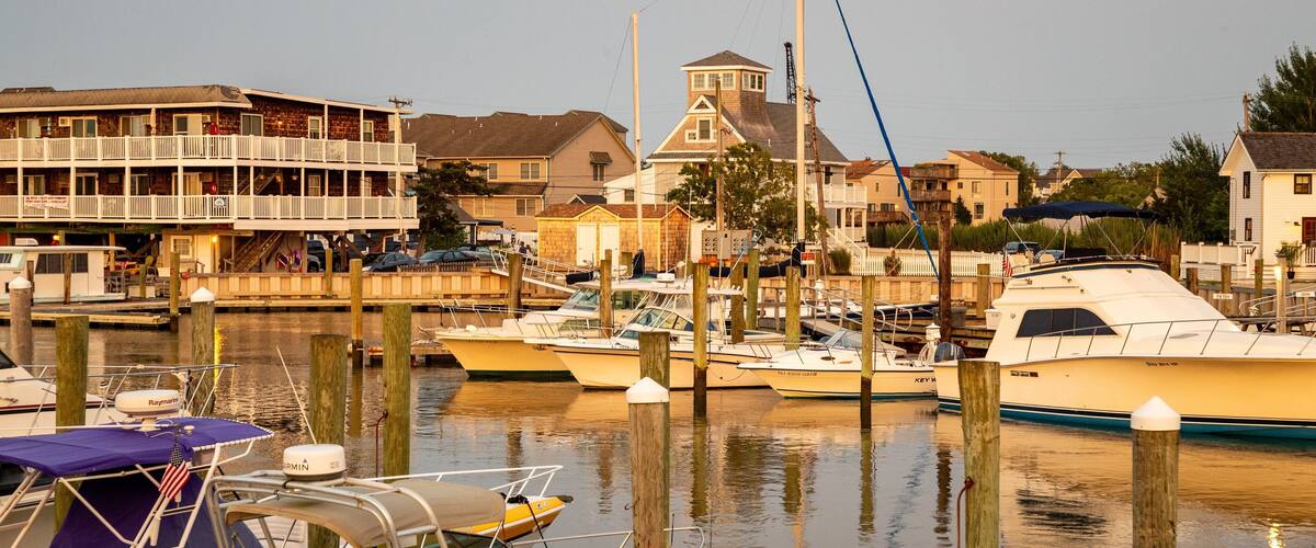 A marina with several boats docked, a wooden pier, and residential buildings in the background.
