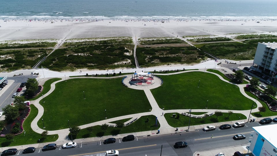 A coastal cityscape with a large sandy beach, a central green park with a circular design, and a parking area with cars.