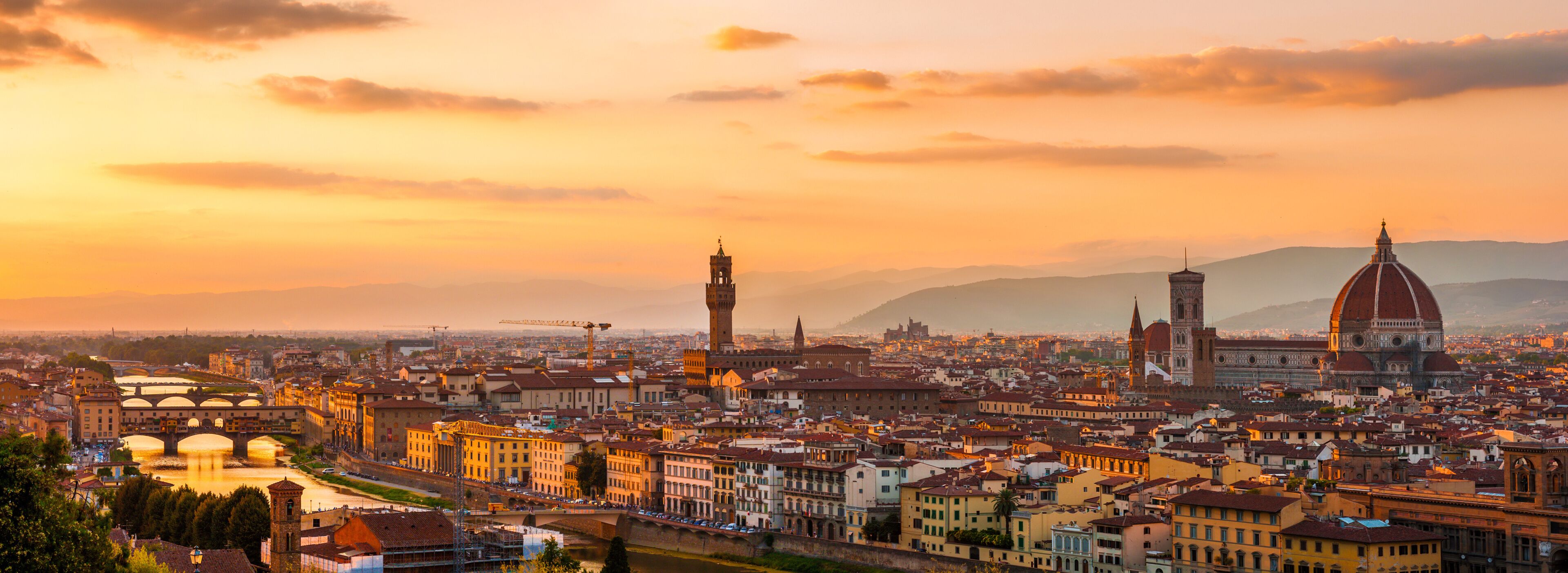 A panoramic view of a city with prominent historical architecture, including a large dome and bell towers, during sunset.
