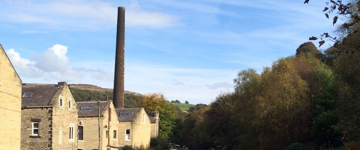 A calm river with a stone bridge, a tall chimney, and buildings along the riverbank.