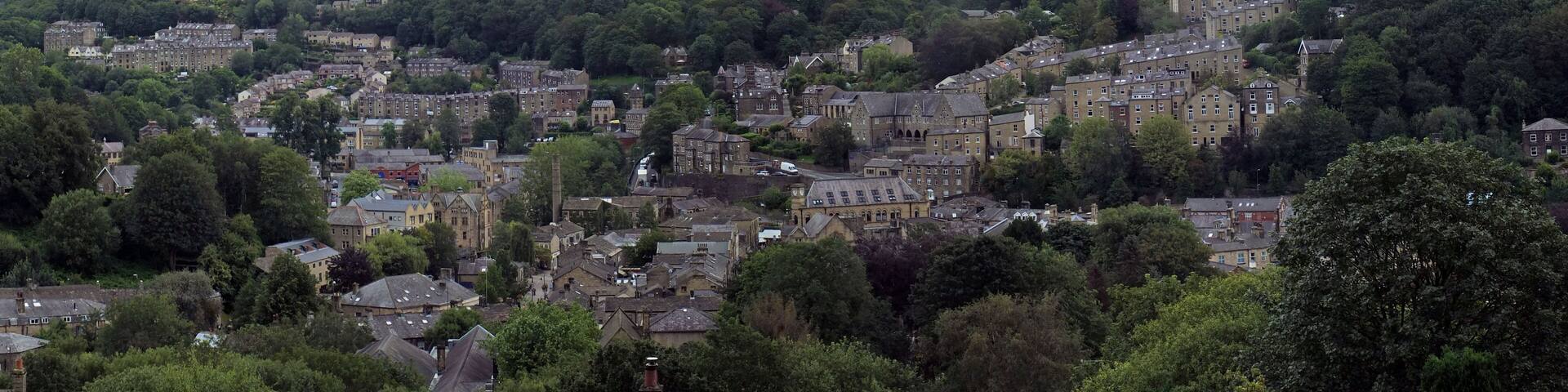 a wide panoramic view of the town of hebden bridge with hillside streets surrounded by trees and pennine fields