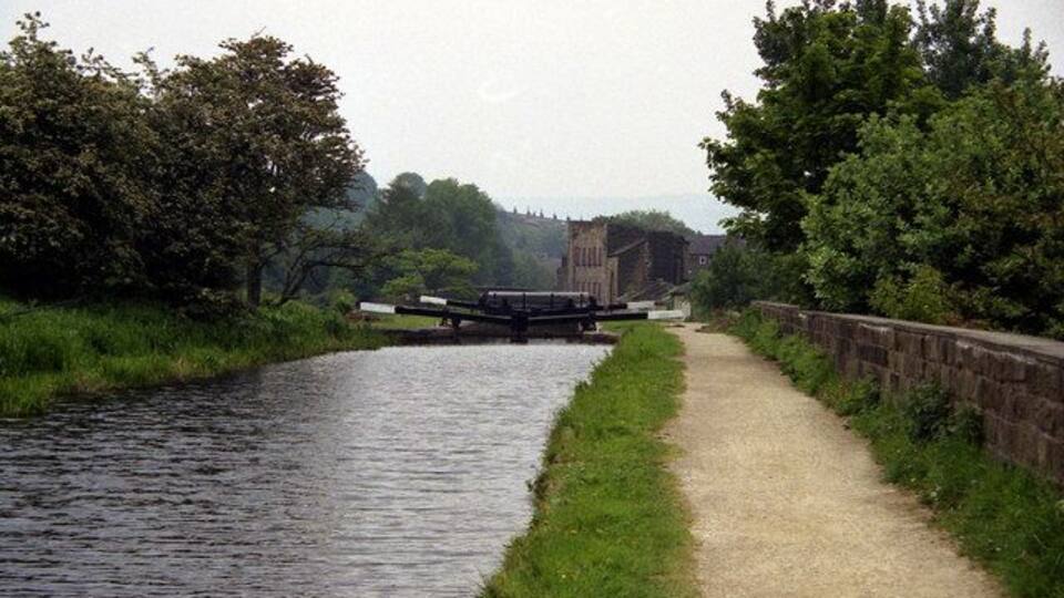 A narrow waterway flanked by grassy banks and trees, with a stone wall on one side and a path on the other.