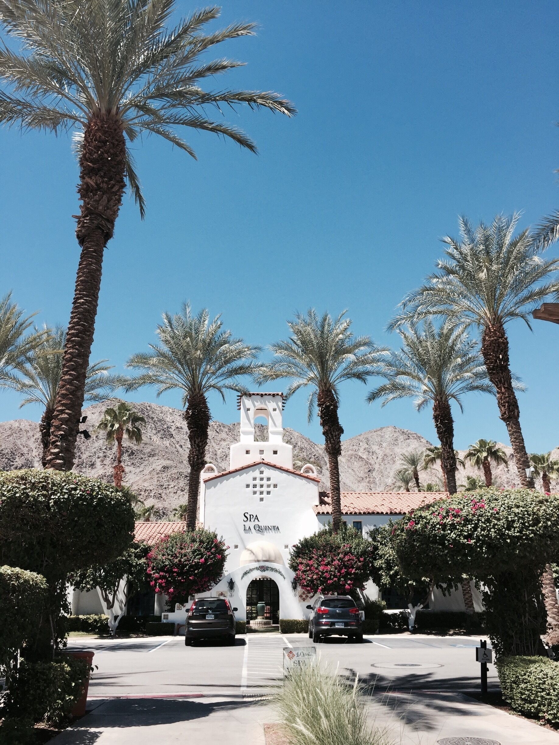 A white building with a bell tower labeled "SPA LA QUINTA" surrounded by palm trees and a mountainous backdrop.