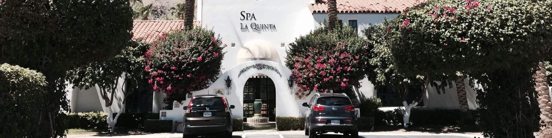 A white building with a bell tower labeled "SPA LA QUINTA" surrounded by palm trees and a mountainous backdrop.