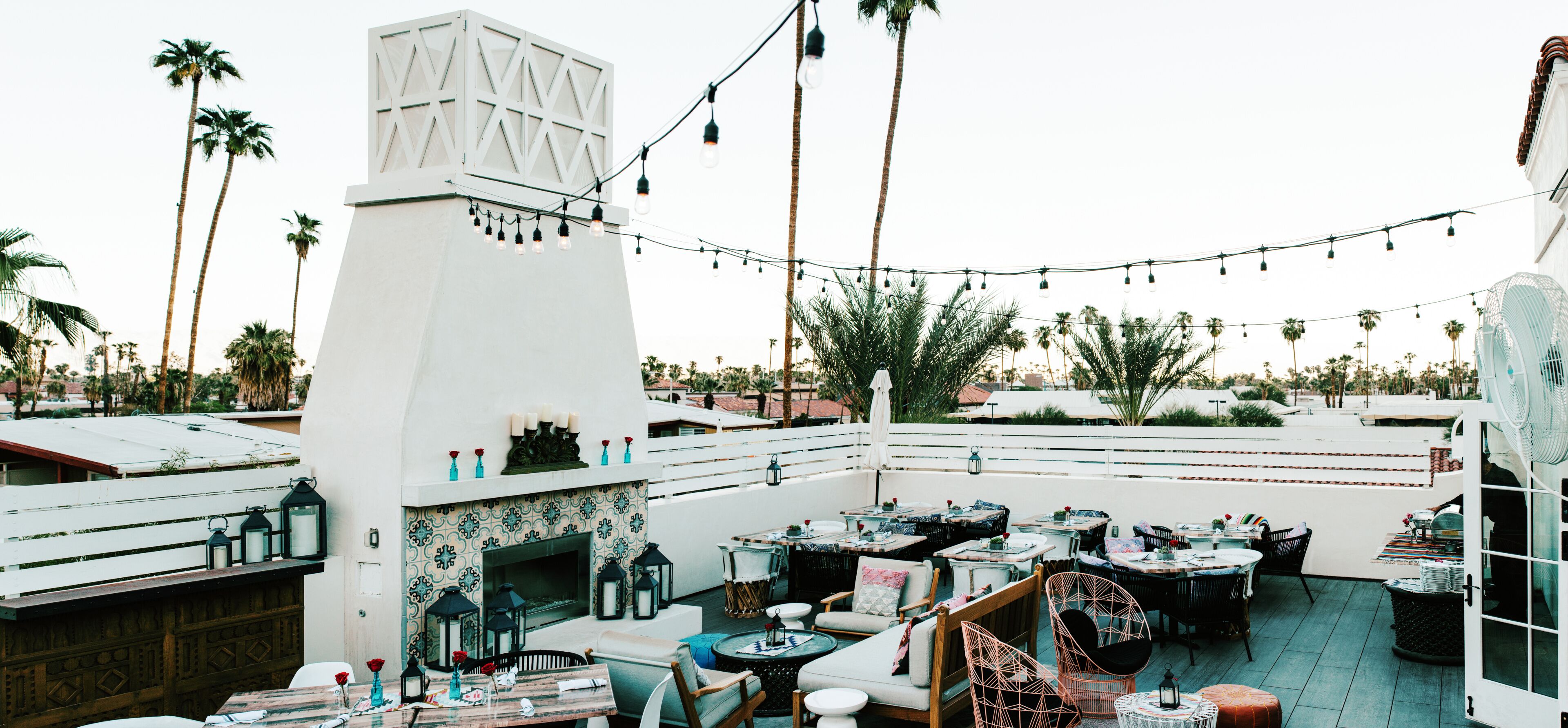 An outdoor patio with string lights, tables, chairs, a fireplace, and palm trees.