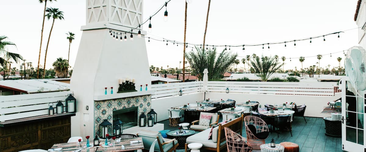 An outdoor patio with string lights, tables, chairs, a fireplace, and palm trees.