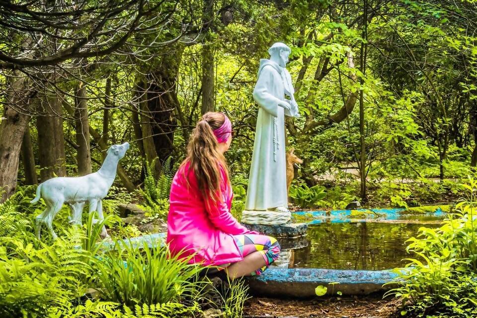 A person sitting by a pond with statues and a dog in a lush green forest.