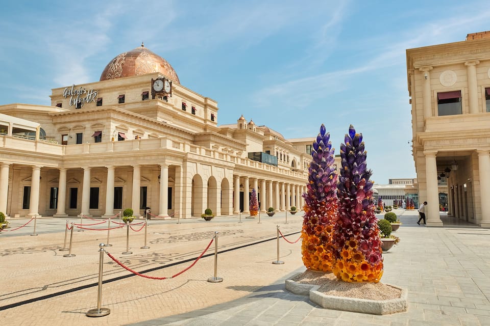 Un imposant bâtiment doté d’un dôme et de colonnes, entouré d’une place où se trouvent une sculpture de verre et des cordes rouges.