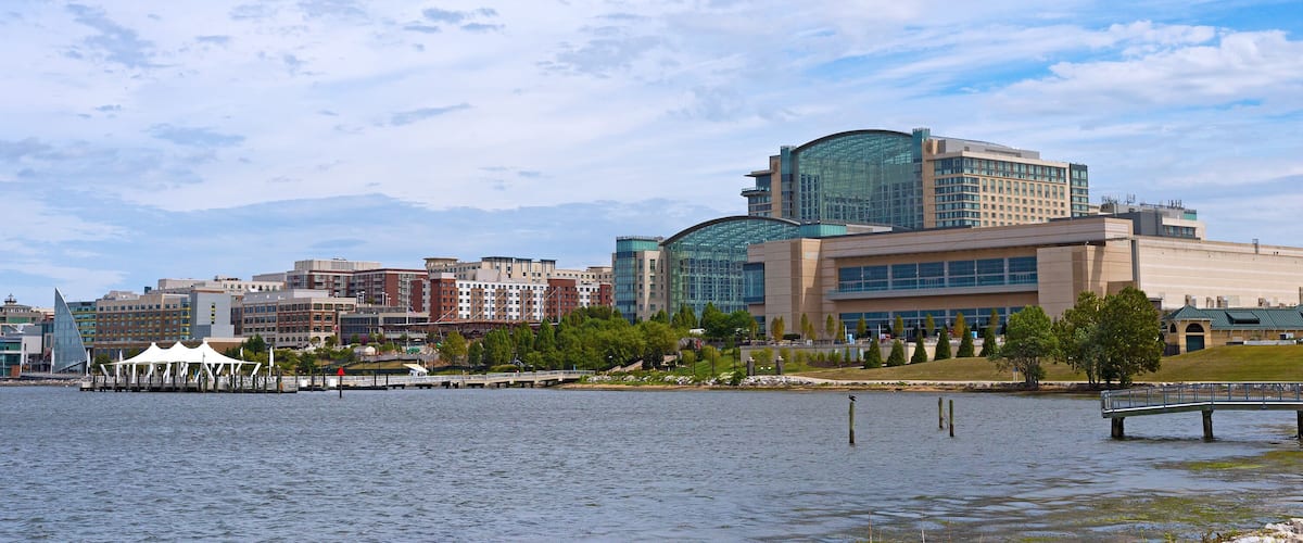 National Harbor waterfront panorama in Oxon Hill, Maryland, USA. Sun shines through cumulus clouds on National Harbor pier and modern buildings along coastline of Potomac River.