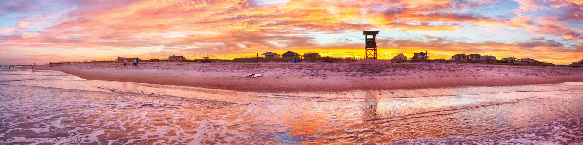 A beach at sunset with a lifeguard tower, houses, and people on the sand.