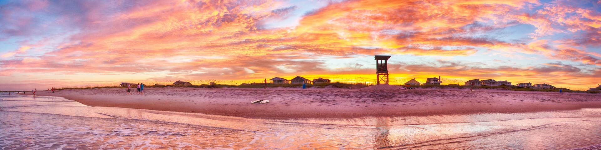 Uma praia ao pôr do sol, com uma torre salva-vidas, casas e pessoas na areia.