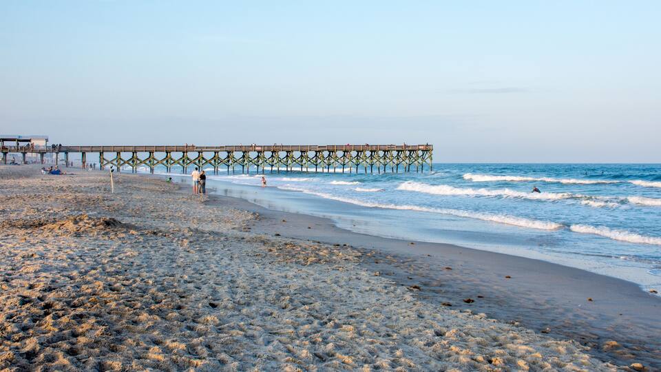 A beach with a wooden pier extending into the ocean, sandy shore, and waves crashing onto the beach.