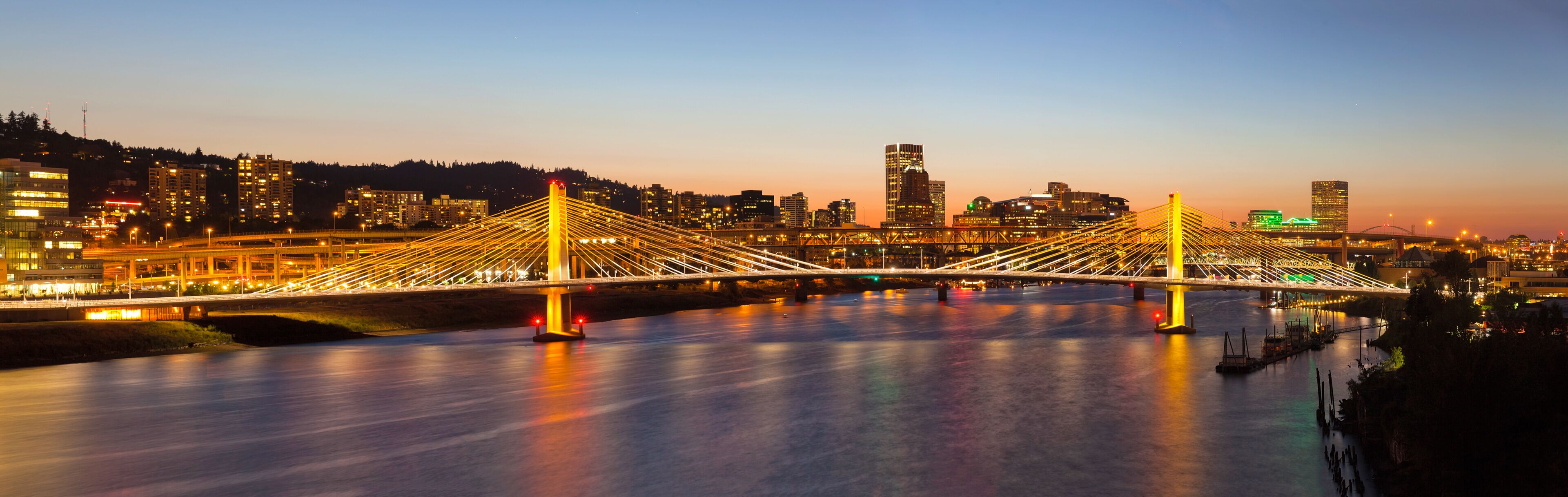 A modern bridge with illuminated cables, a river below, and a city skyline in the background.