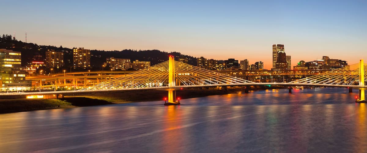 A modern bridge with illuminated cables, a river below, and a city skyline in the background.