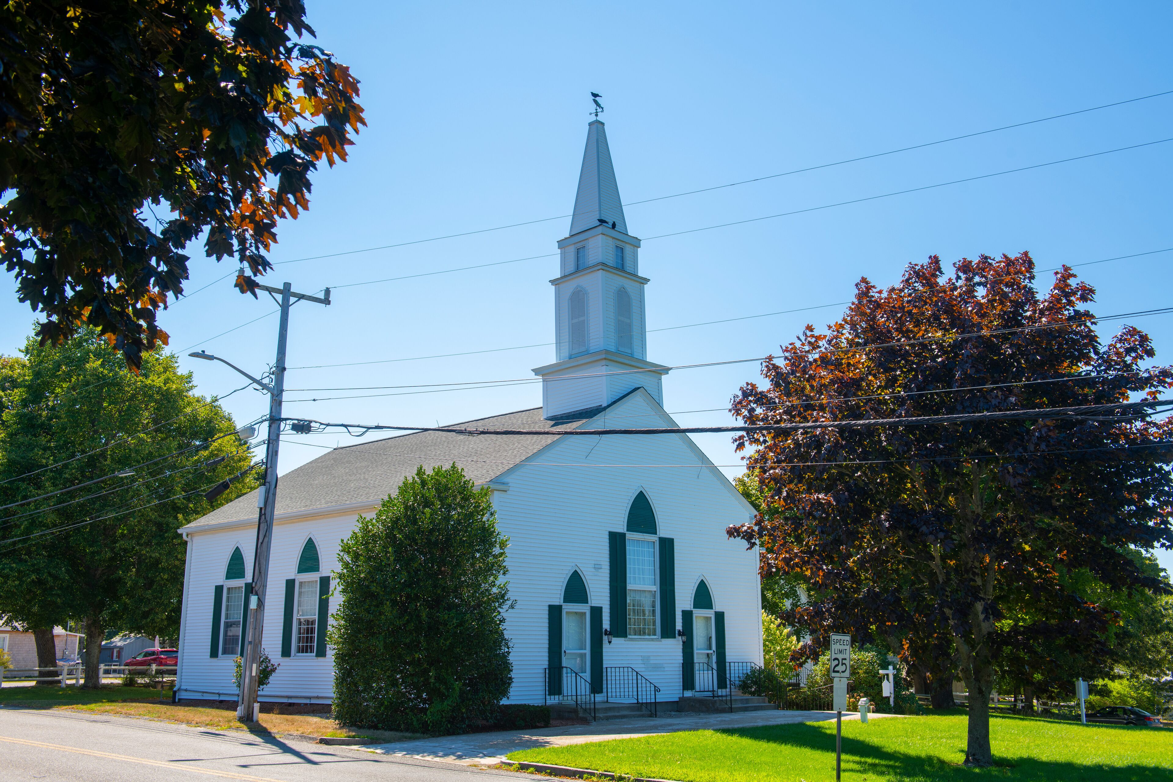 A white church with a steeple and green shutters, surrounded by trees and a clear sky.