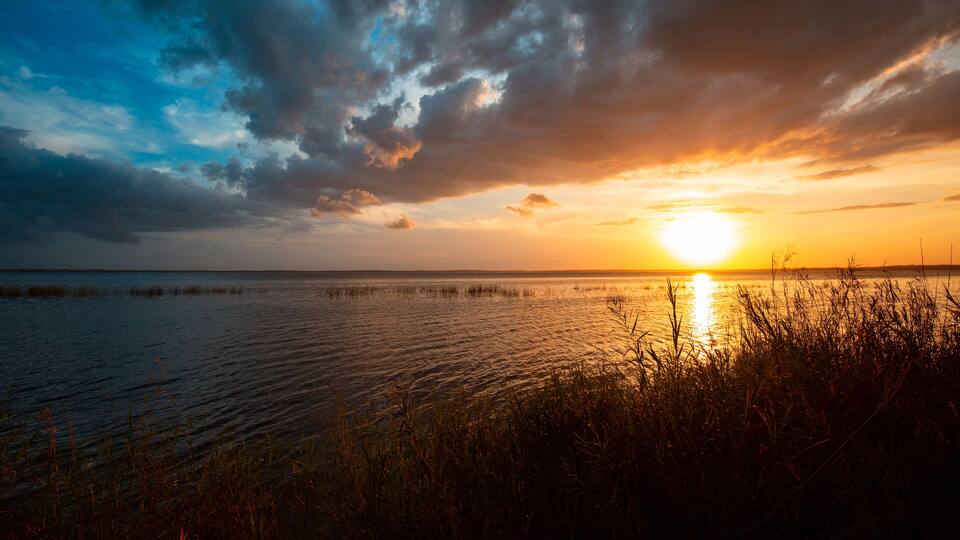 A sunset over a calm water body with a grassy foreground and a cloudy sky.