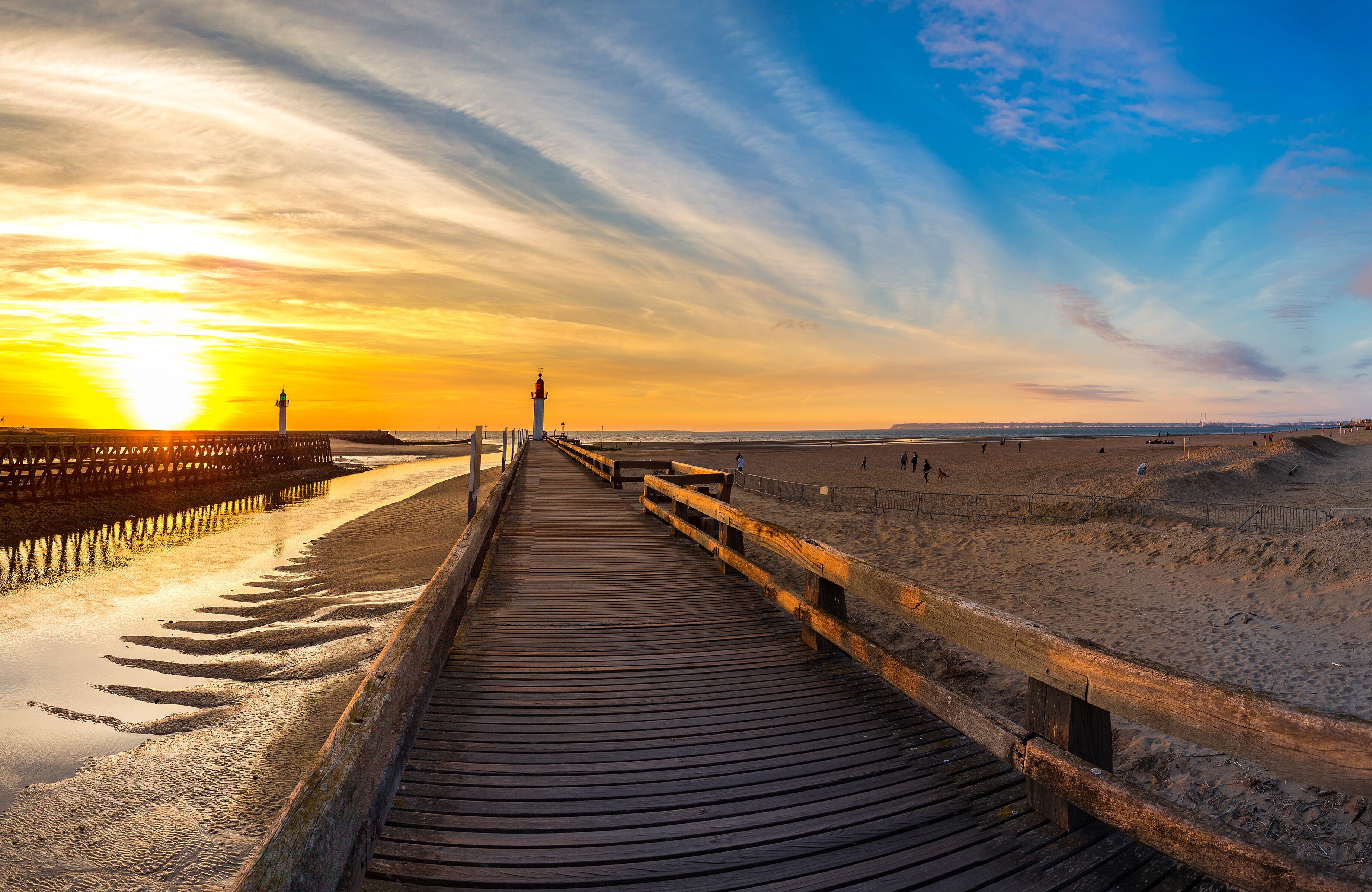 A wooden boardwalk leading to a lighthouse, with a sunset and beach in the background.