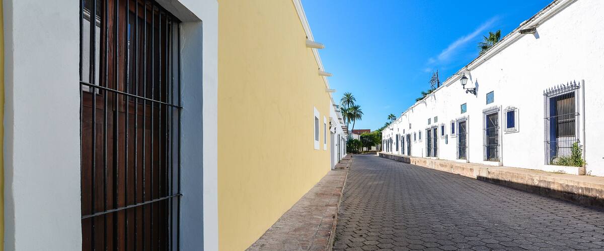 A cobblestone street flanked by white and yellow buildings with barred windows and doors.