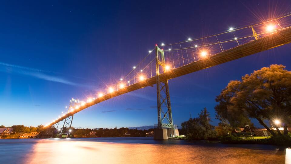 A nighttime view of a large suspension bridge with lights, spanning over a body of water, with trees and buildings in the background.