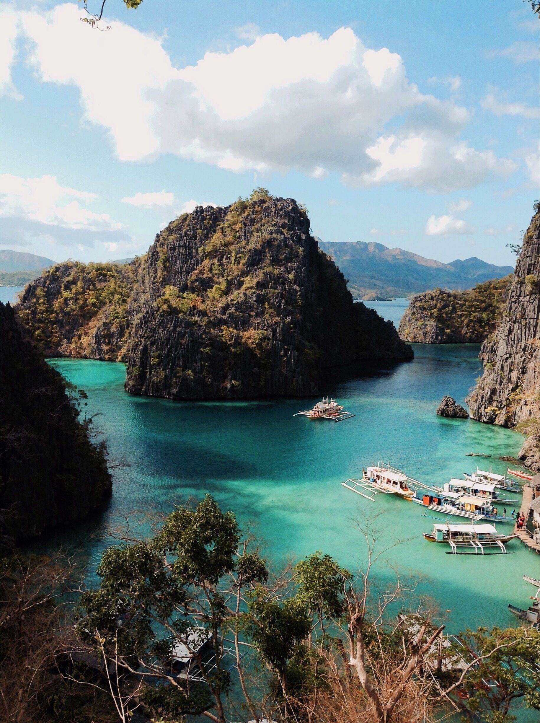 A coastal landscape with a prominent rocky island surrounded by turquoise waters, several boats docked near the shore, and lush greenery on the island.