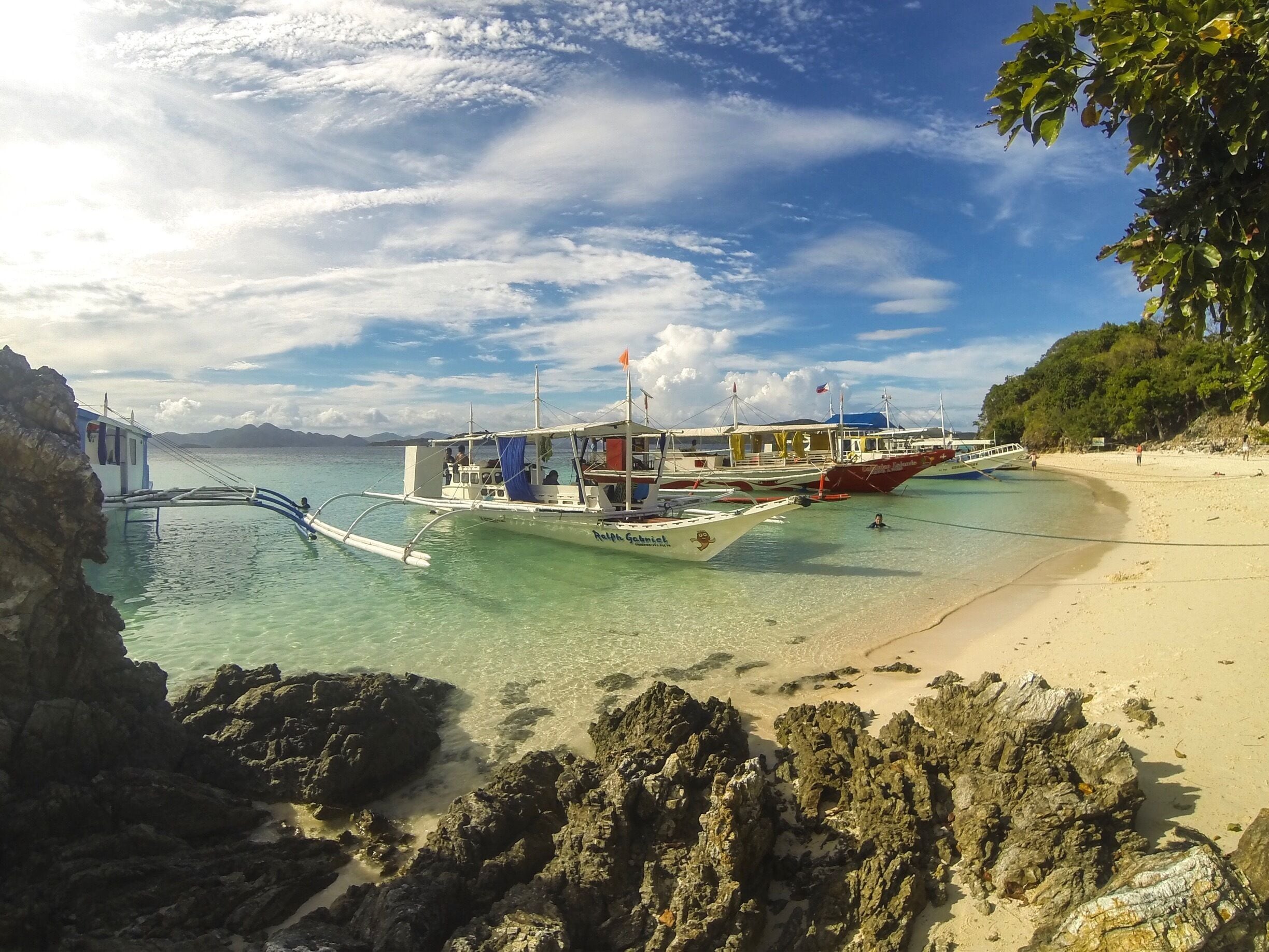 A beach with clear turquoise water, anchored boats, and rocky formations.