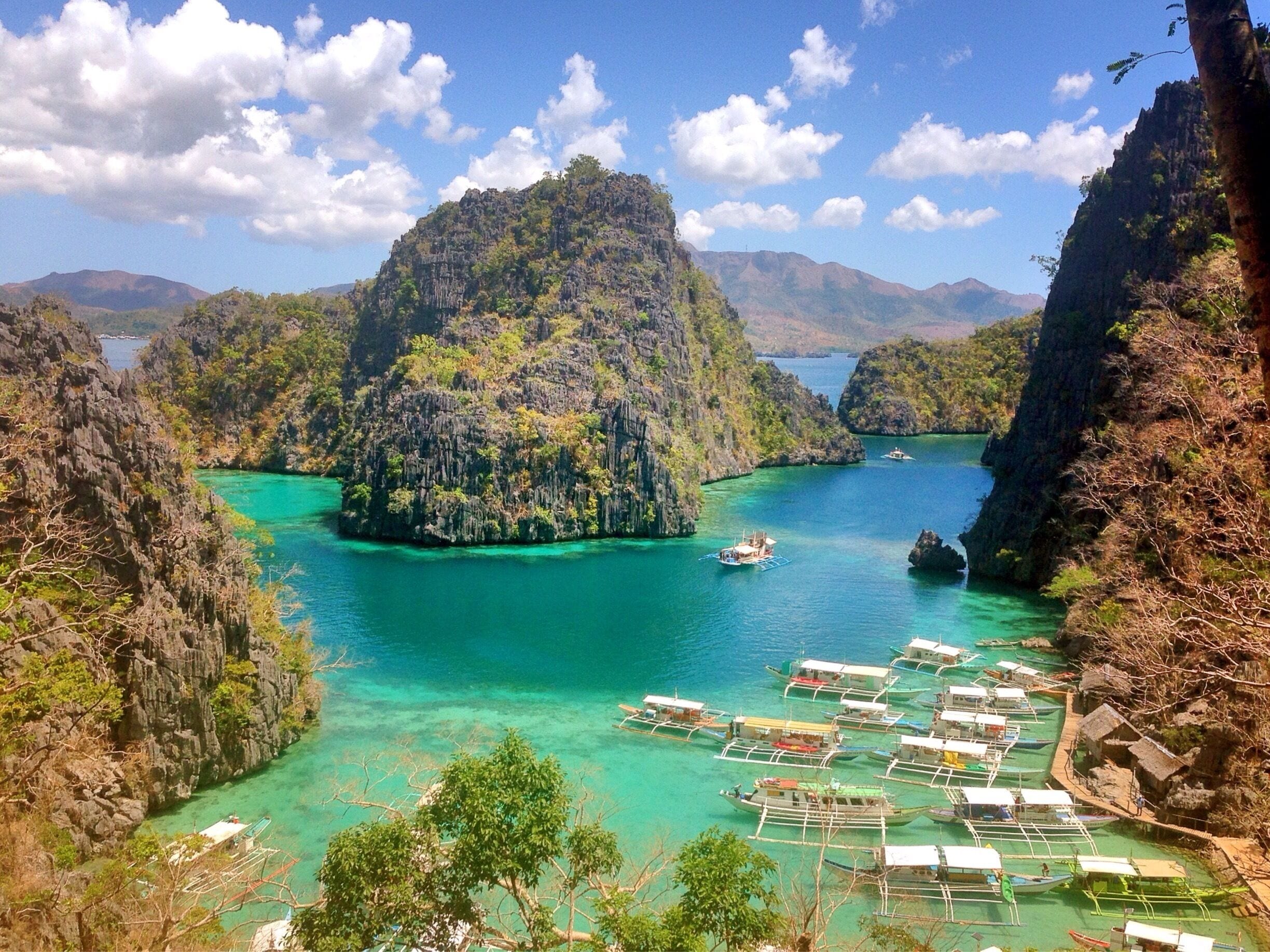 A coastal landscape with clear turquoise waters, a large island with dense vegetation, and several boats anchored near the shore.