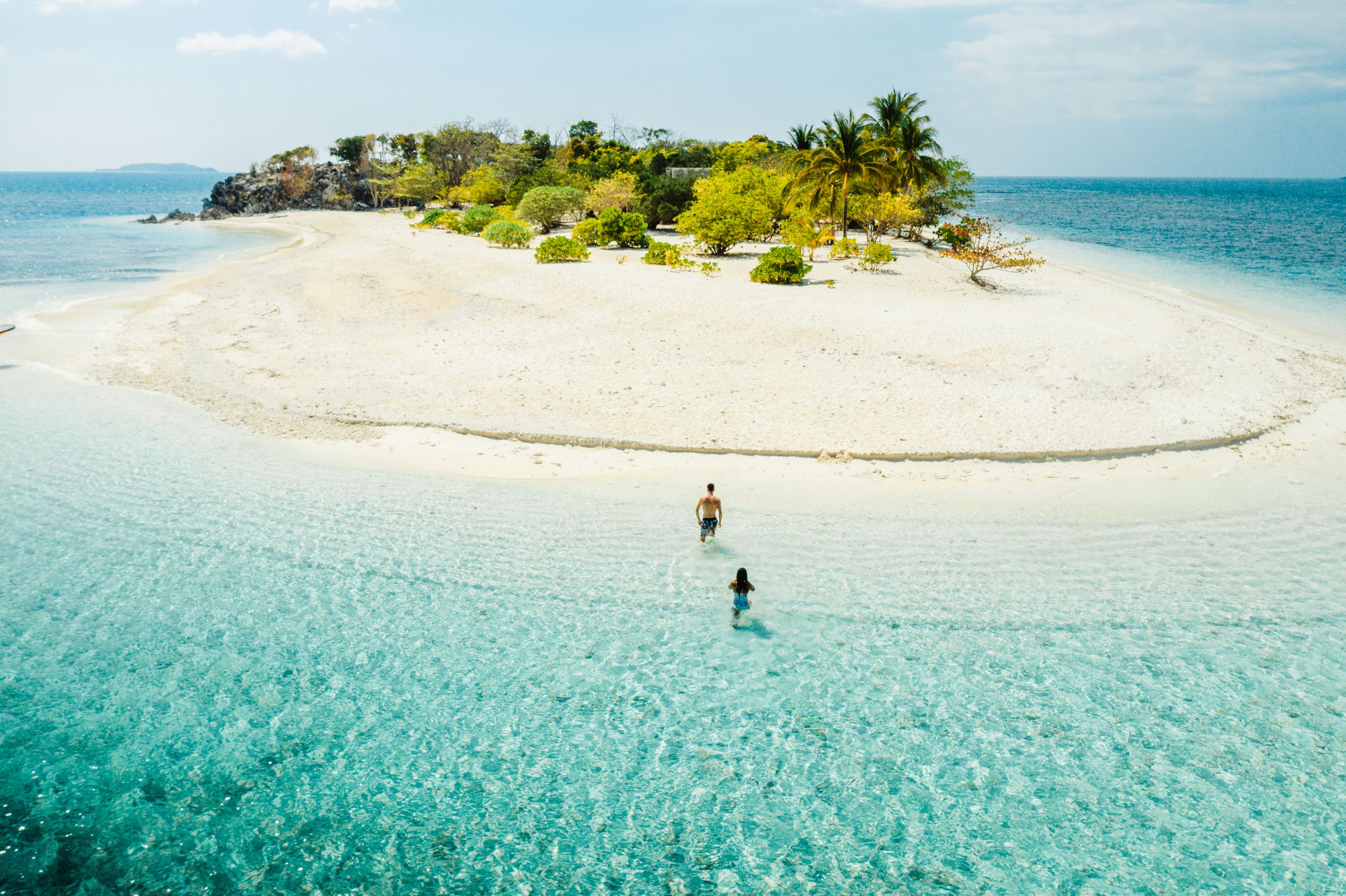 Una playa arenosa con agua turquesa cristalina, una pequeña isla con vegetación y dos personas caminando hacia el agua.