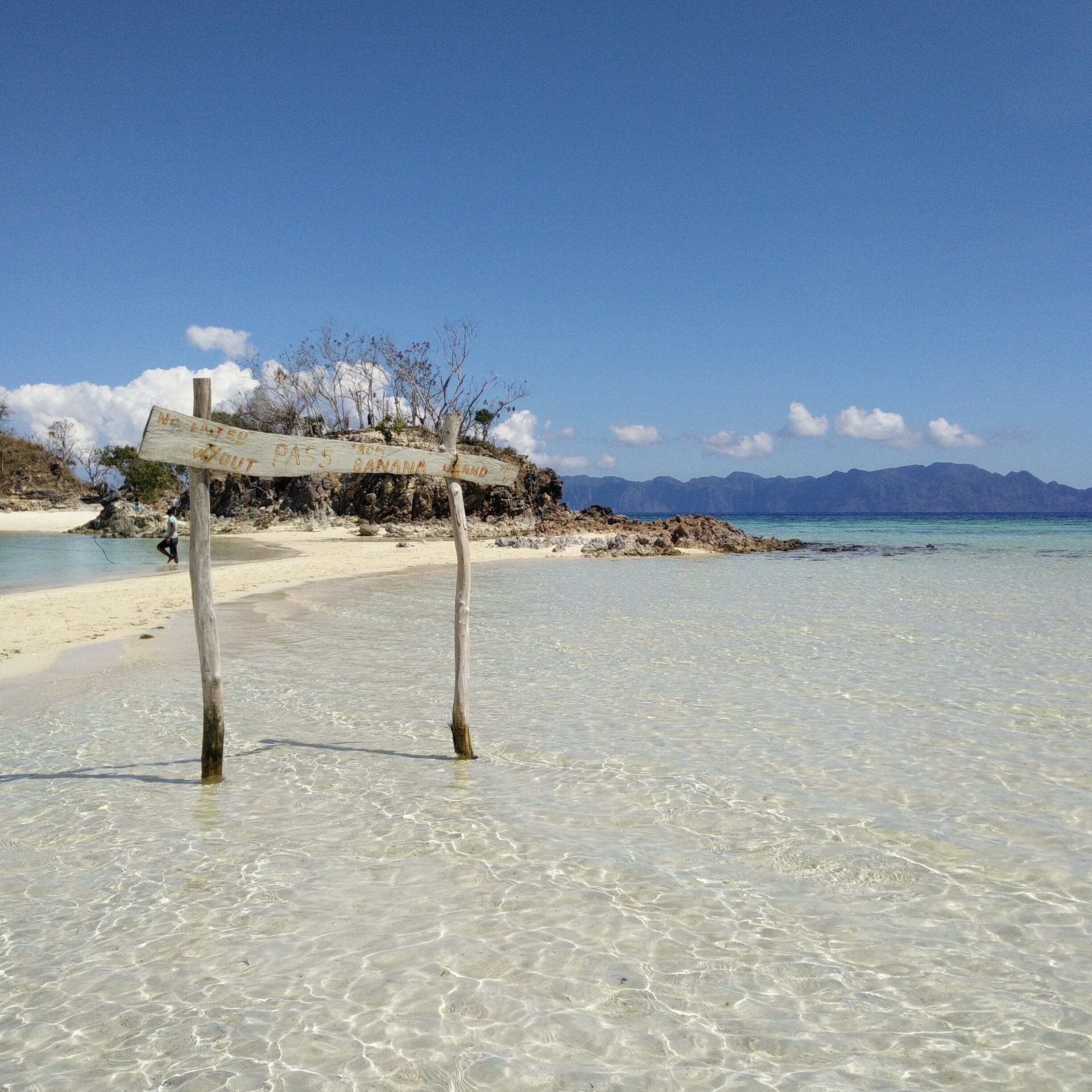 A beach with clear shallow waters, a wooden signpost, and a distant mountain range.