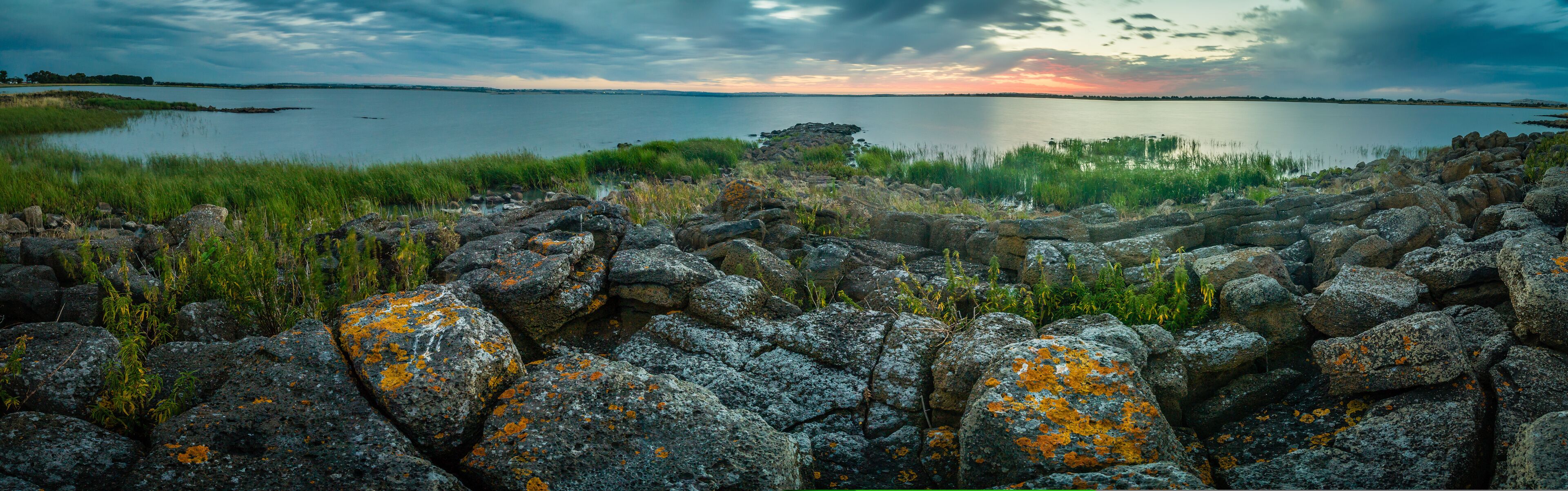 A rocky shoreline with lichen-covered rocks and a calm lake at sunset.
