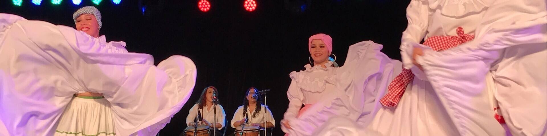 A group of dancers in white costumes performing on a stage with musical instruments and lighting.
