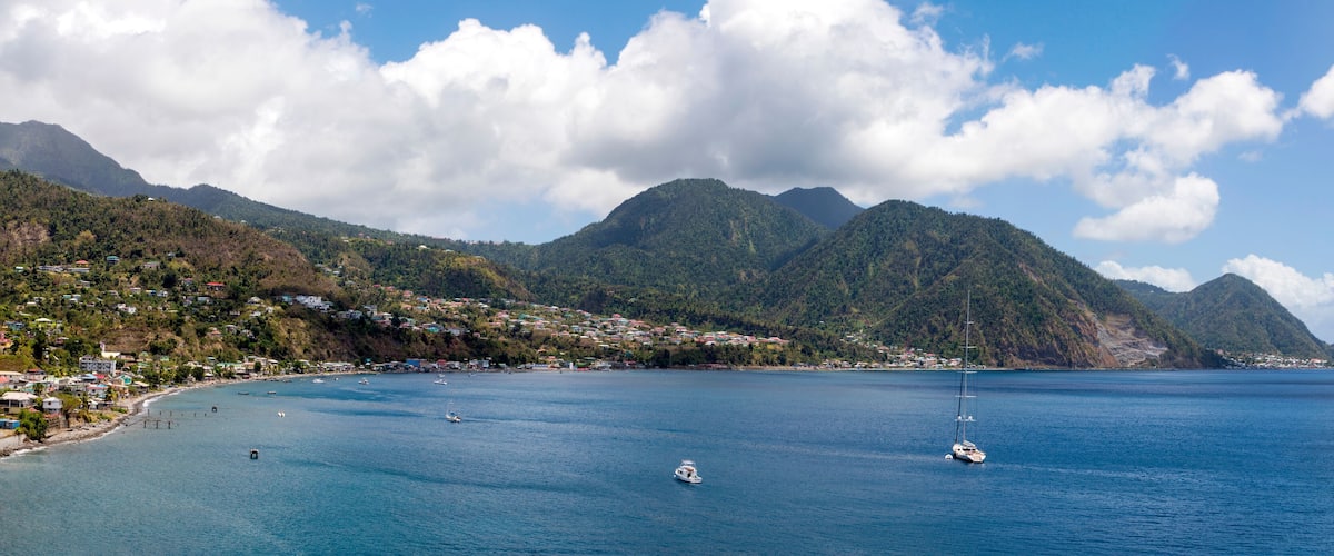 A coastal town with a clear blue sea, boats, and lush green mountains in the background.