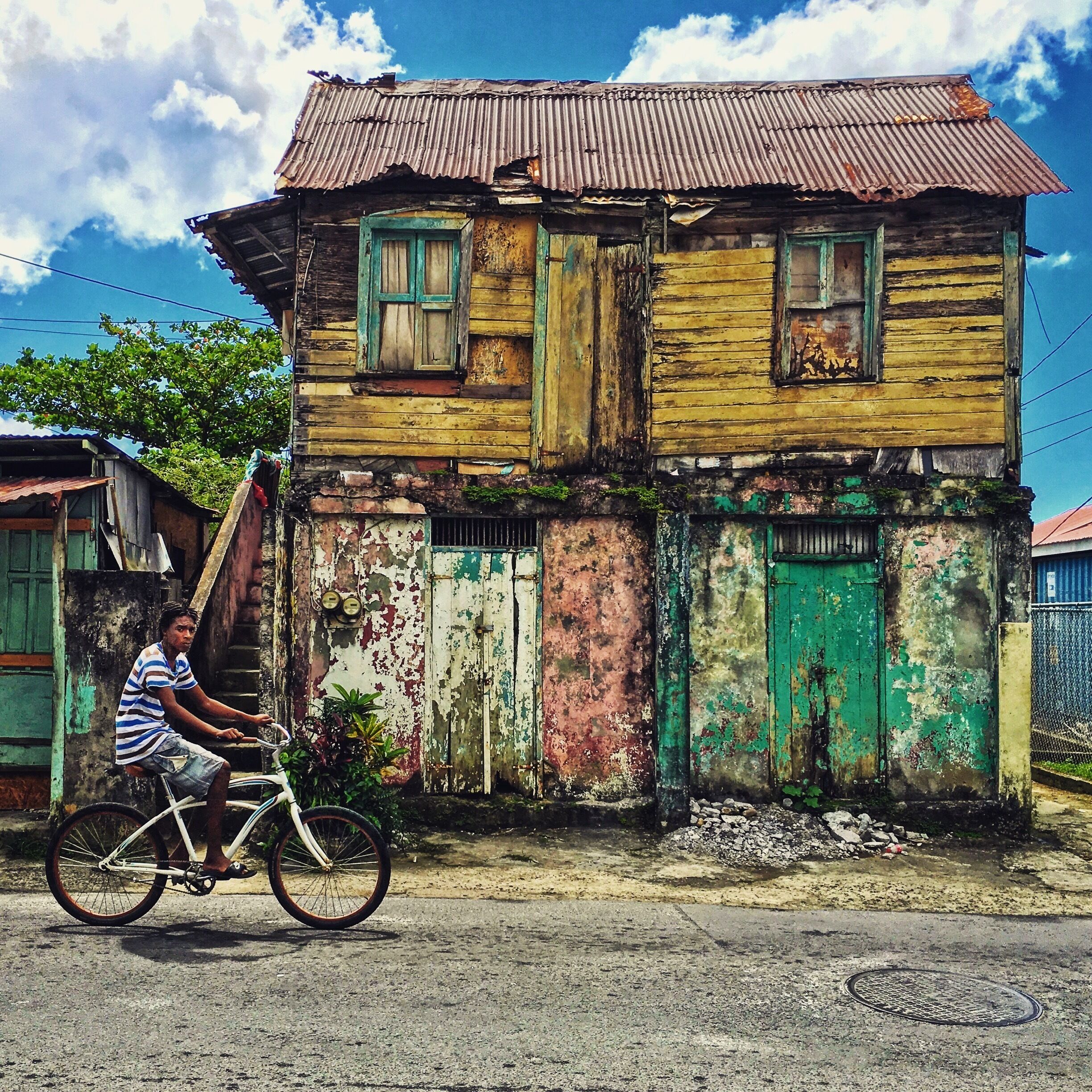 A dilapidated wooden house with a corrugated metal roof, a man riding a bicycle, and a clear blue sky with clouds.