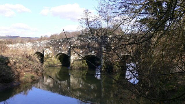 A stone bridge with three arches over a calm river, surrounded by trees and open fields.