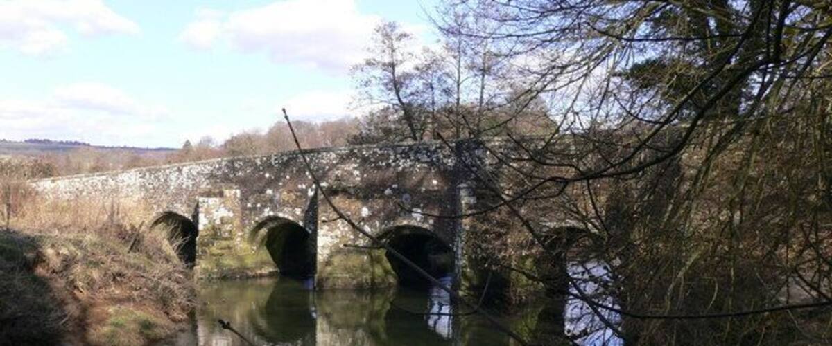 A stone bridge with three arches over a calm river, surrounded by trees and open fields.