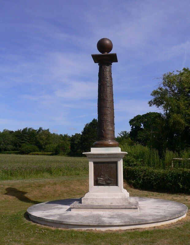 Um monumento alto e cilíndrico com uma cúpula esférica no topo, rodeado por uma base circular e uma placa.