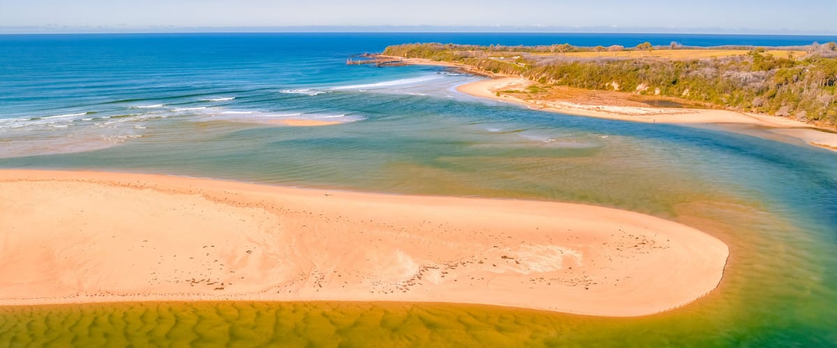 A coastal landscape with clear blue waters, a sandy beach, and a distant shoreline with vegetation.