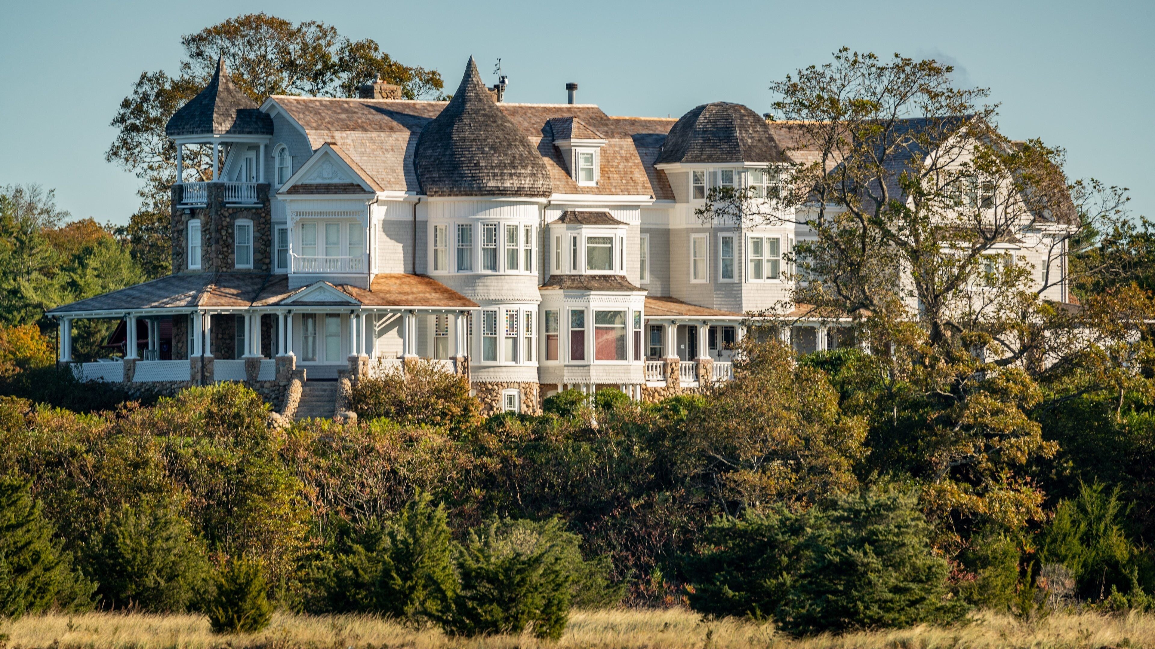 A large, multi-story house with a wrap-around porch, surrounded by dense trees and a grassy field.